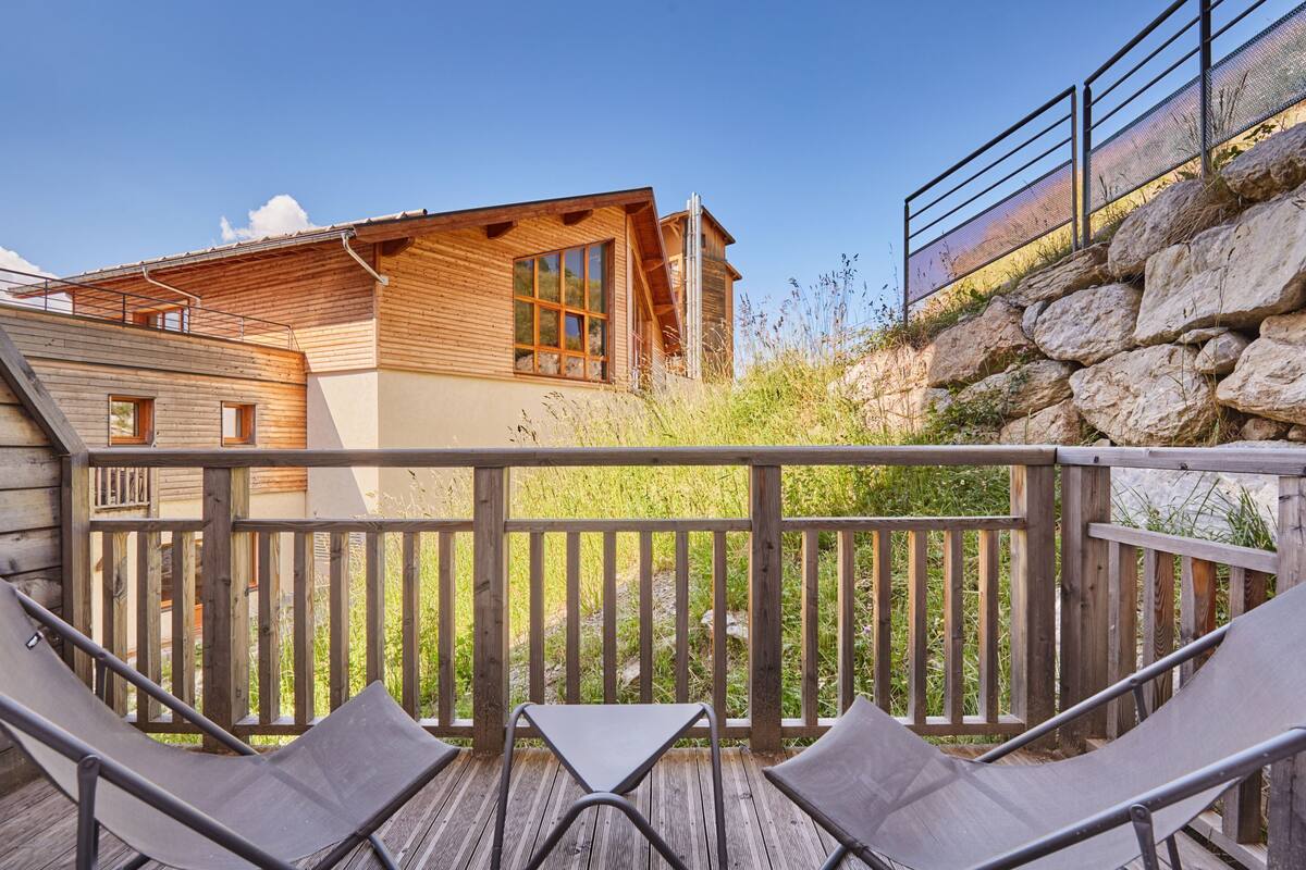 A wooden deck with two chairs and a table, overlooking a building with large windows and a stone wall.