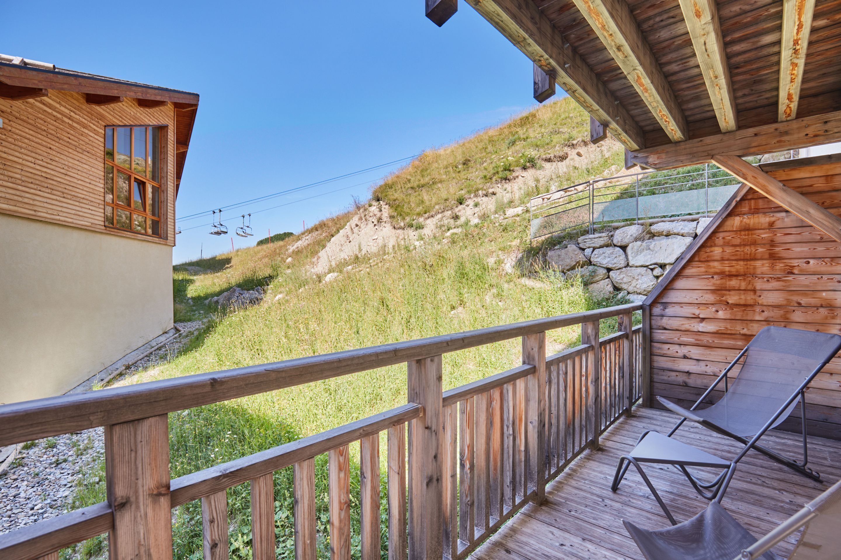 A wooden balcony with a view of a grassy hillside and a stone wall.