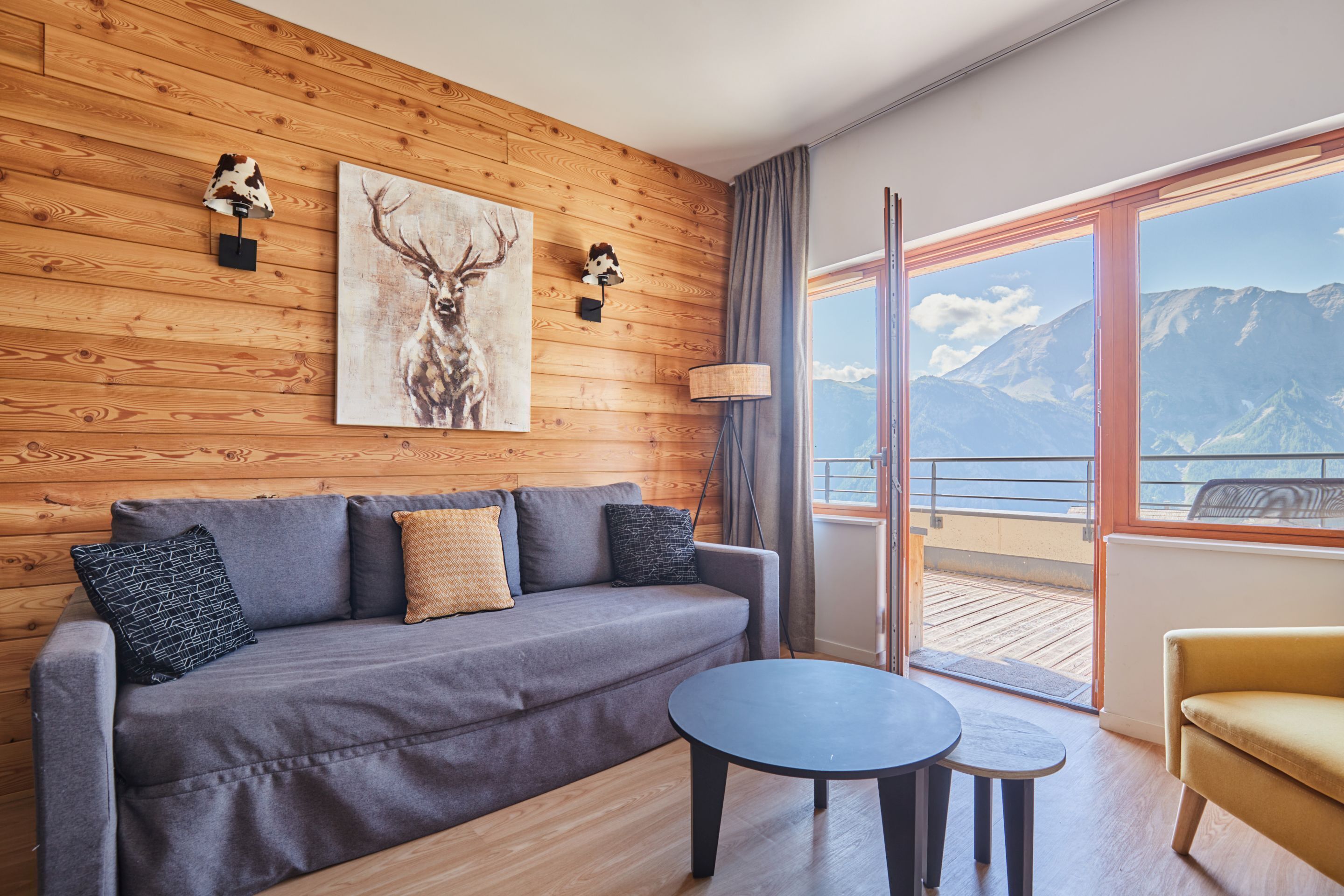 Une pièce moderne avec un mur en bois, un canapé gris, une petite table ronde et une vue sur les montagnes par de grandes fenêtres.