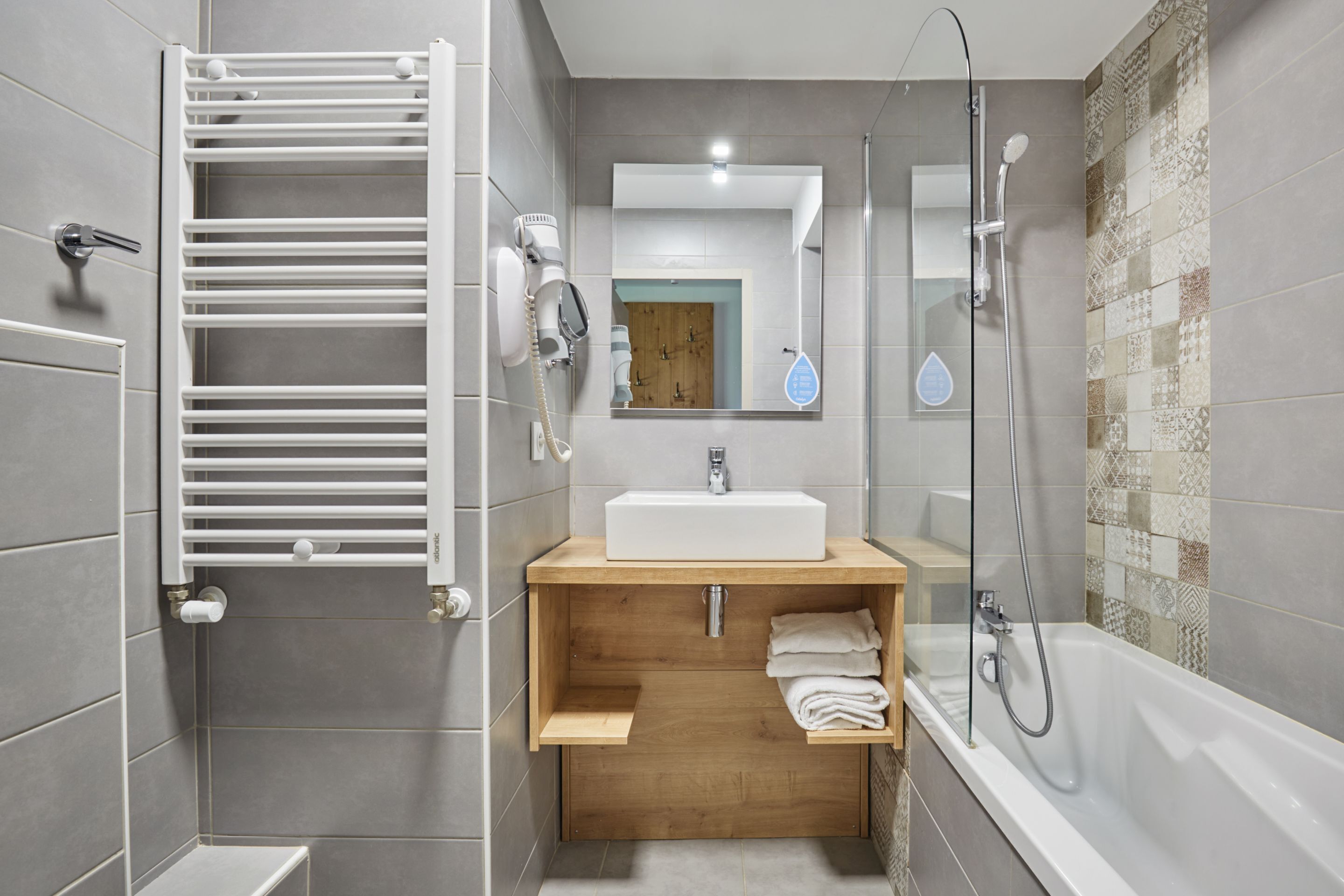 Een moderne badkamer met een witte wasbak, houten wastafelmeubel, handdoekrek en een douchegedeelte.
