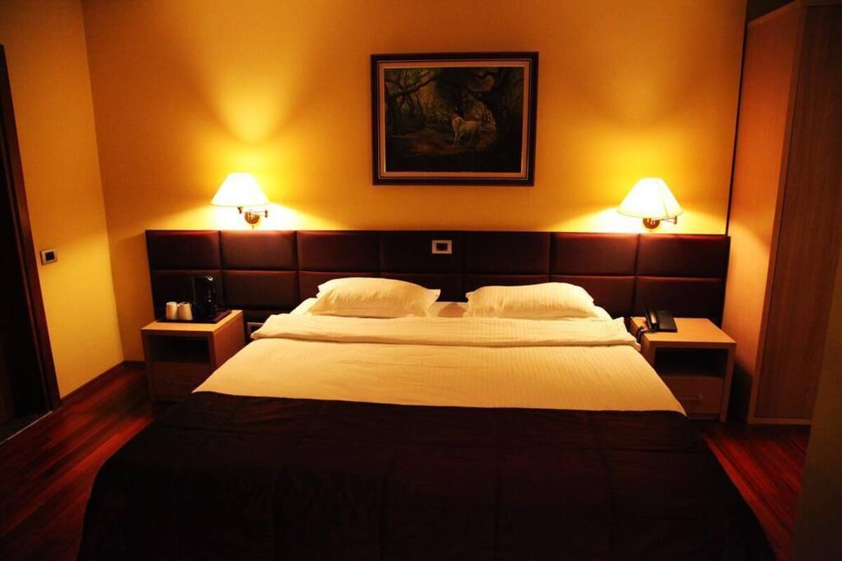 A hotel room with a large bed, two bedside tables, a nightstand, a painting on the wall, and two wall-mounted lamps.