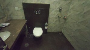 A small, dimly lit bathroom with a white toilet, a dark door, and a tiled wall. - Eden Green (Ganaur)