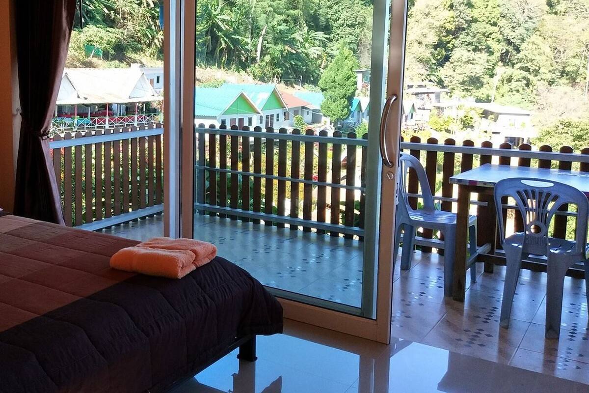 A bedroom with a bed, pillows, and a view of a balcony with a railing and a table.