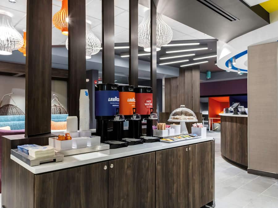Um saguão de hotel moderno, com balcão de recepção, máquinas de café e área de estar com sofá e almofadas.