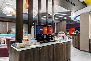 A modern hotel lobby with a reception desk, coffee machines, and a seating area with a sofa and cushions.