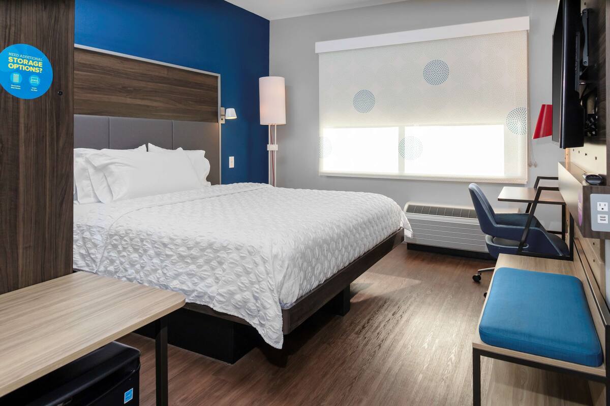 A hotel room with a bed, a desk, a chair, a TV, and a window with blinds.