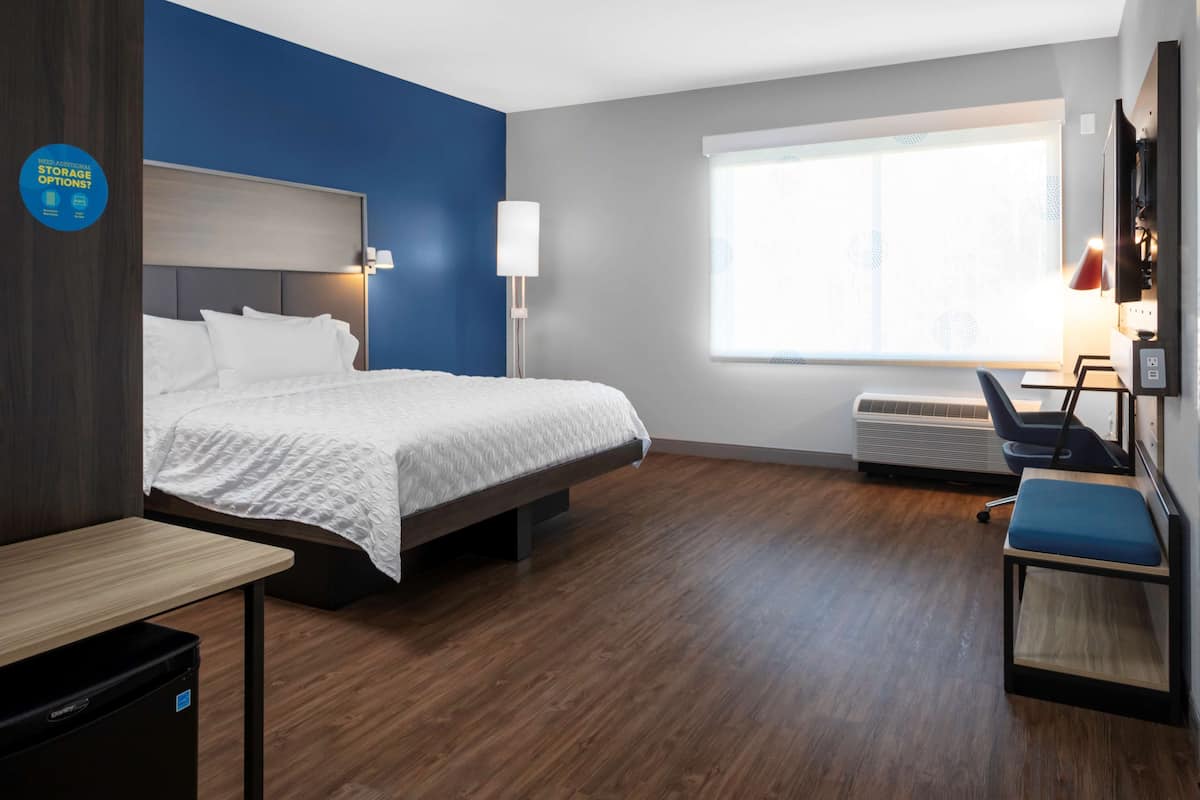 A hotel room with a bed, a desk, a chair, a window, and an air conditioning unit.