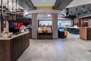 A modern hotel lobby with a central counter, a breakfast area, and a seating area with blue chairs. - TRU by Hilton Lynchburg (Lynchburg)