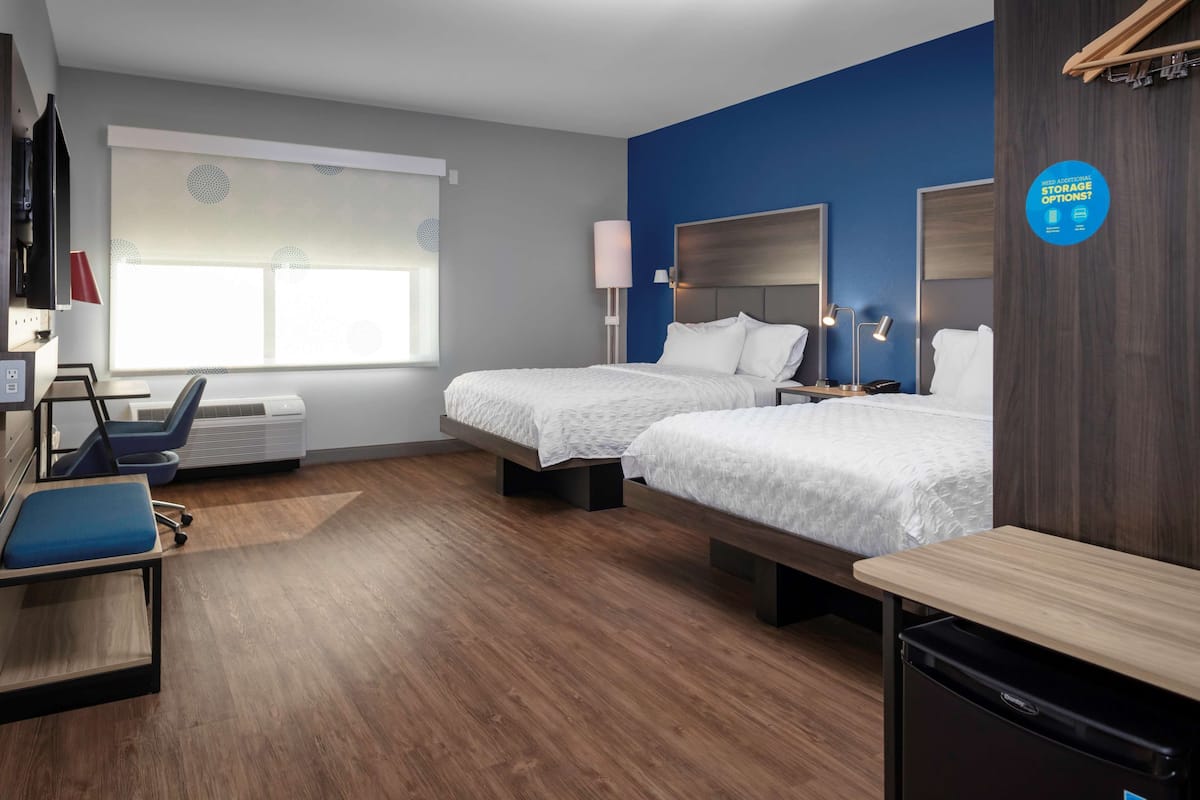 A hotel room with two beds, a desk, a chair, a TV, and a window with blinds.