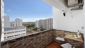 A balcony with a stone wall, a wooden table with chairs, and a view of high-rise buildings. - Holiday Apartment “Da Praia” with Air Conditioning, Balcony, Shared Garden & WiFi (Portimão)
