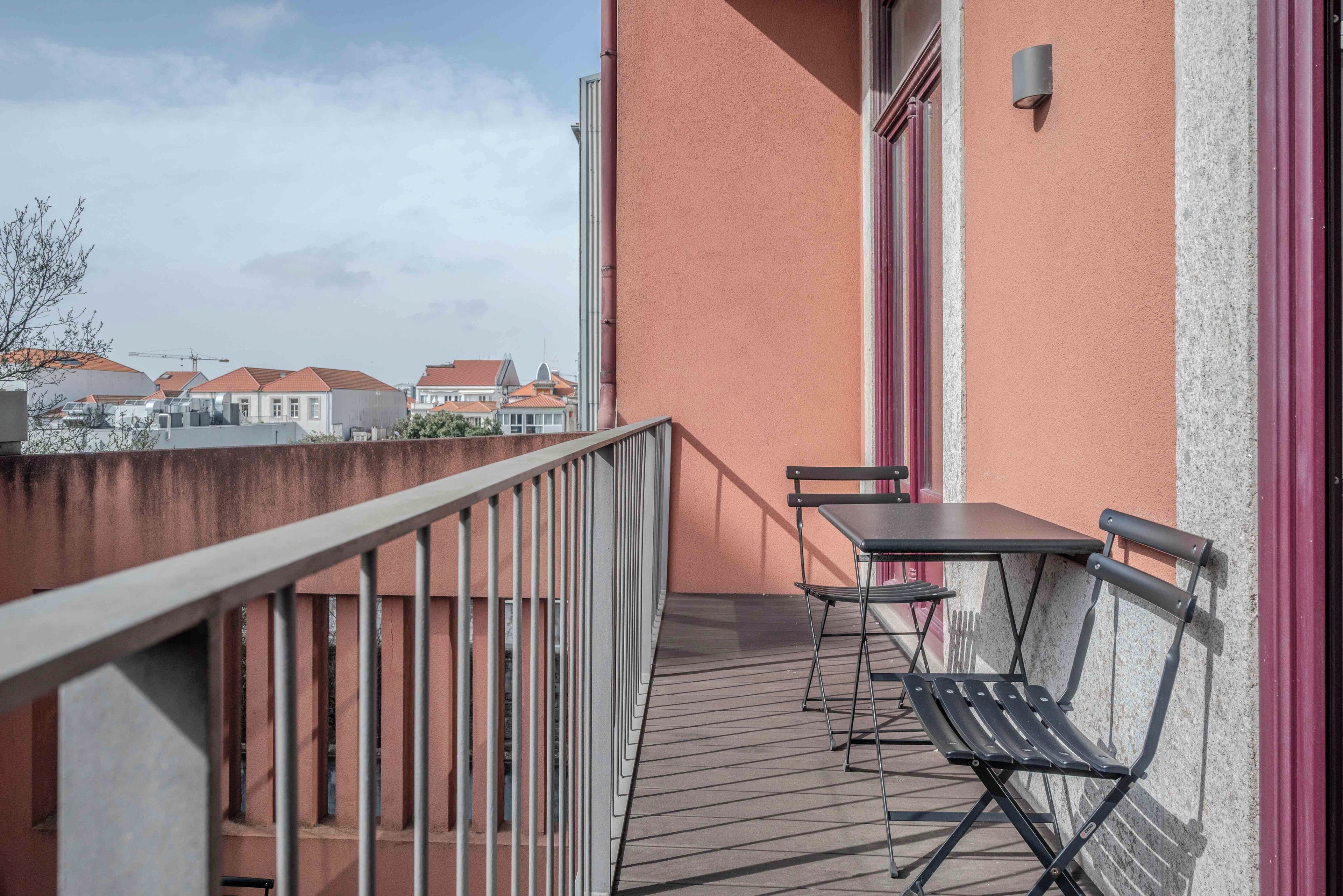 A balcony with a table and chairs, a metal railing, and a view of buildings in the background.