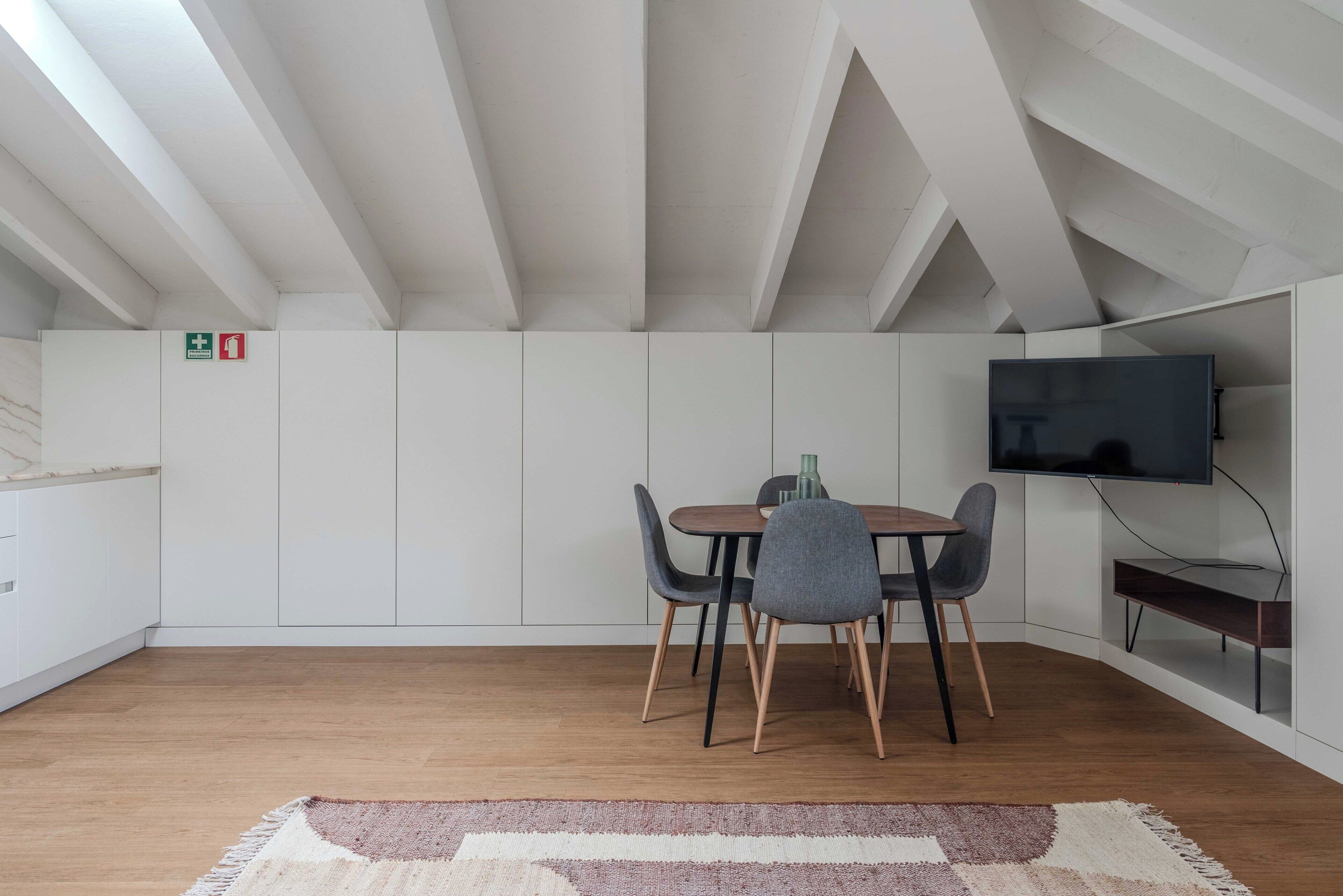 A modern dining area with a wooden table and chairs, a flat-screen TV mounted on the wall, and a white ceiling with exposed beams.
