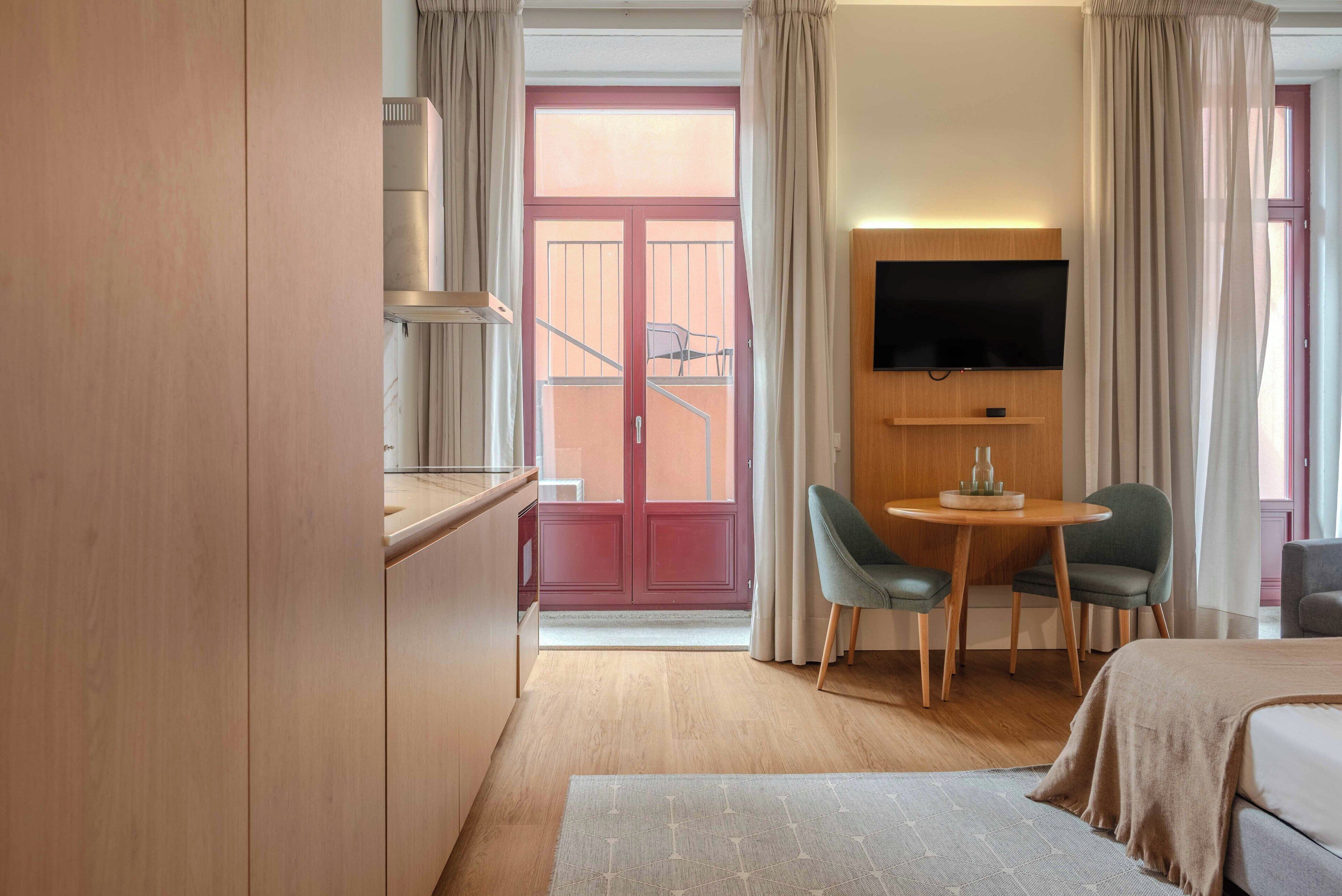 Habitación de hotel con cama, televisor montado en la pared, una pequeña mesa con dos sillas y un balcón con barandilla y una silla.