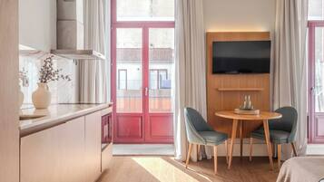 A modern hotel room with a kitchen area, a dining table with chairs, a flat-screen TV mounted on the wall, and a balcony with red doors.