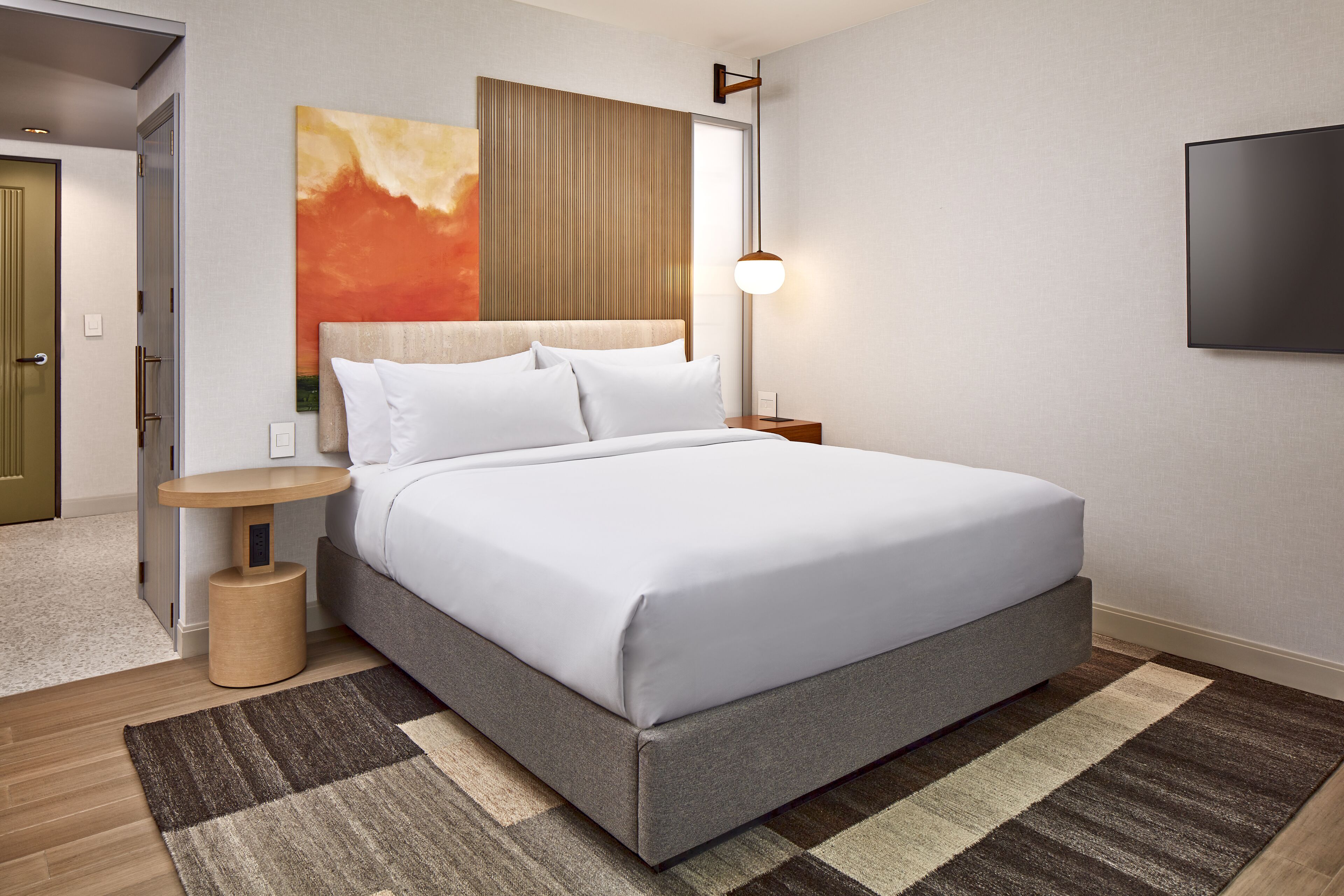 A modern hotel room with a large bed, a bedside table, a wall-mounted TV, and a painting above the bed.