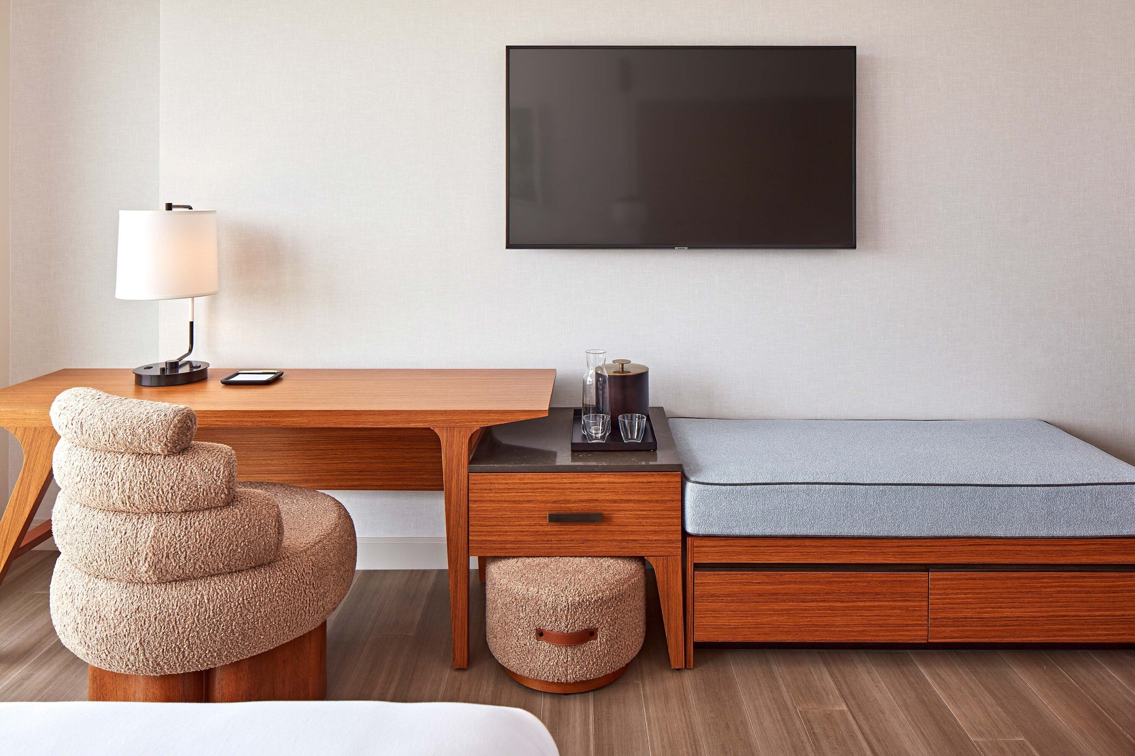 Une chambre d’hôtel compacte, dotée d’un bureau en bois, d’un fauteuil confortable, d’un lit avec un matelas gris, d’un téléviseur fixé au mur et d’une lampe sur le bureau.