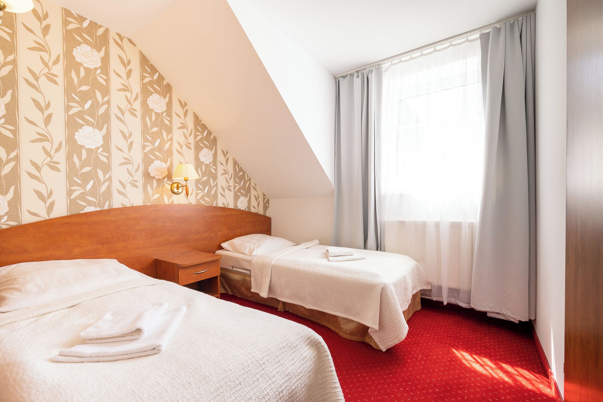 A hotel room with two beds, a wooden headboard, a nightstand, a lamp, and a window with curtains.