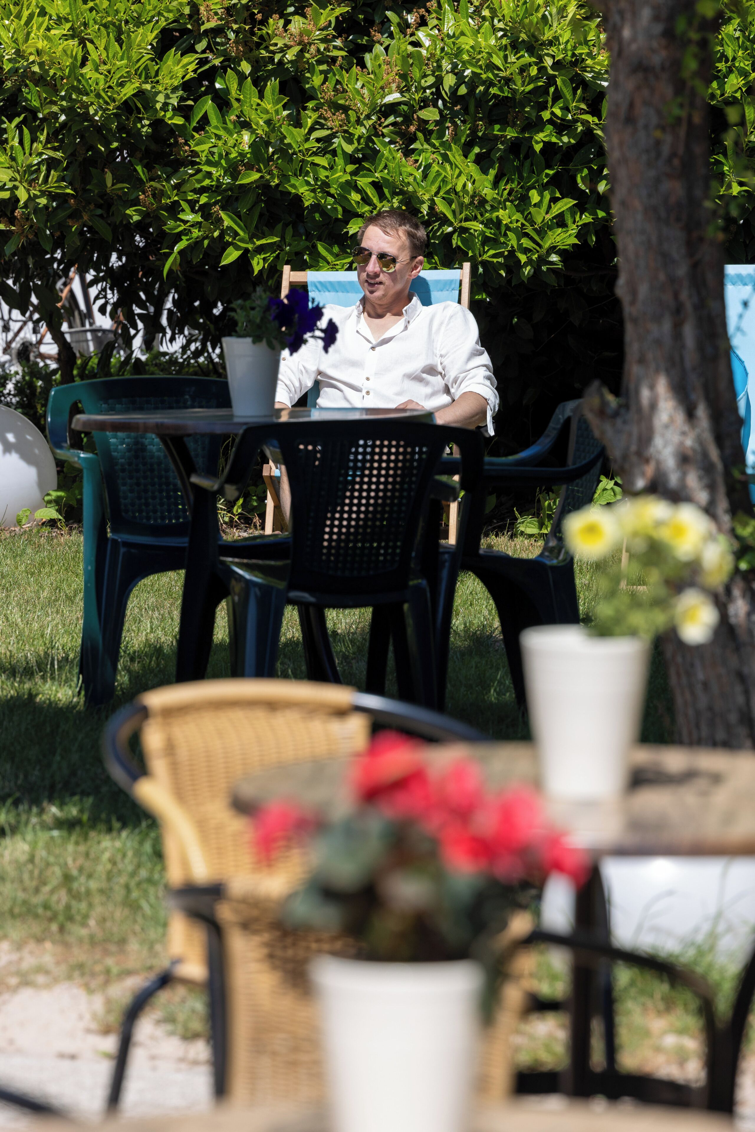 Une personne est assise à une table à l’extérieur, entourée de chaises, avec une plante en pot et un arbre en arrière-plan.