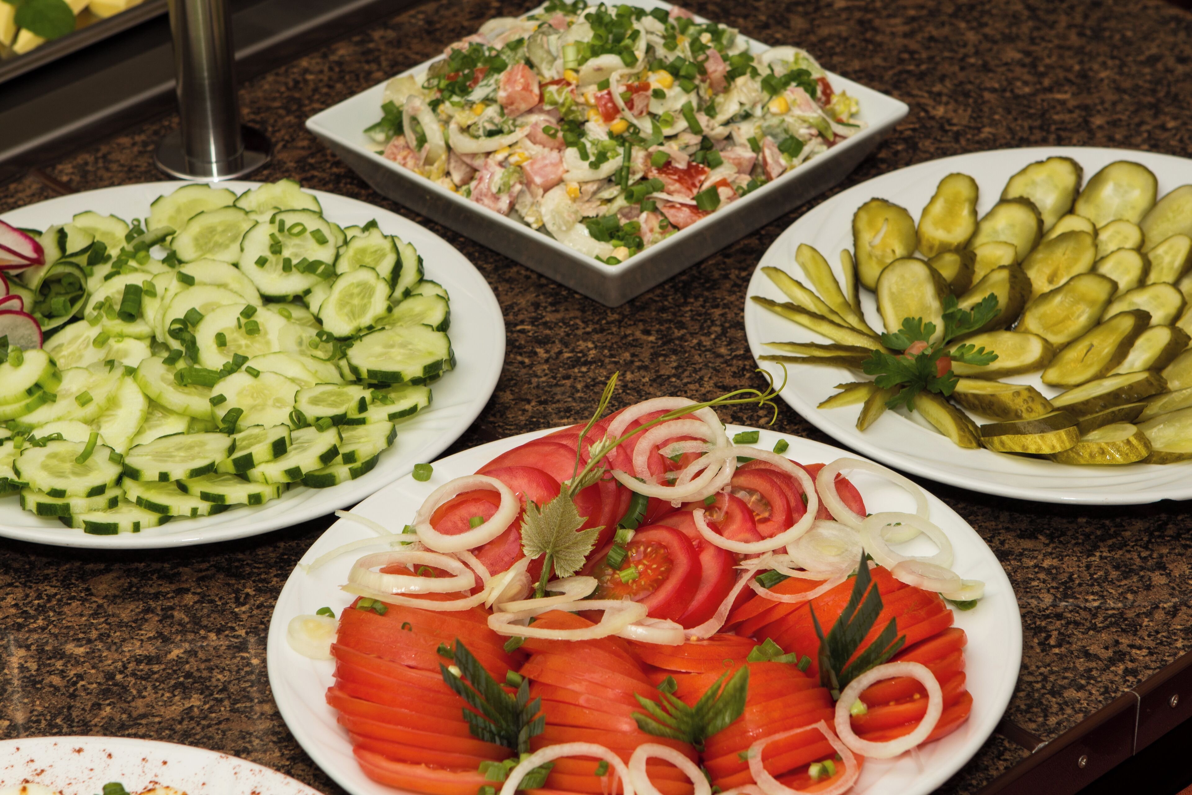 Un buffet proposant divers plats, notamment des tranches de concombre, une salade, des cornichons et une assiette de tomates en tranches, garnie d’oignons.