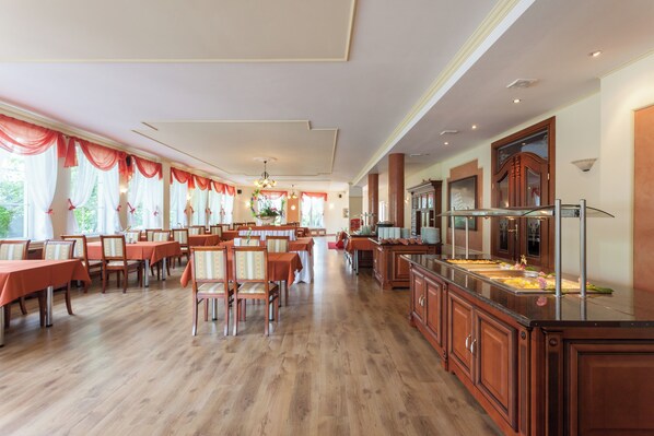 A spacious dining area with multiple tables and chairs, a buffet station, and a view of greenery outside.