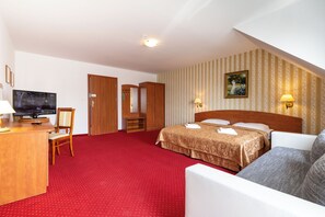 A hotel room with a red carpet, a bed with a headboard, a desk with a television, a chair, and a wardrobe.