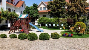 Un terrain de jeux avec un toboggan, entouré d’arbustes soigneusement taillés et comprenant un espace de repos avec des tables et des chaises.