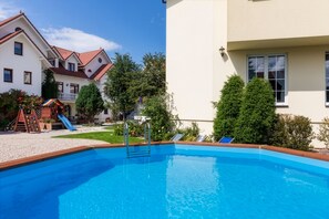 A swimming pool with a wooden deck, a children's play area, and a white building with a red roof.