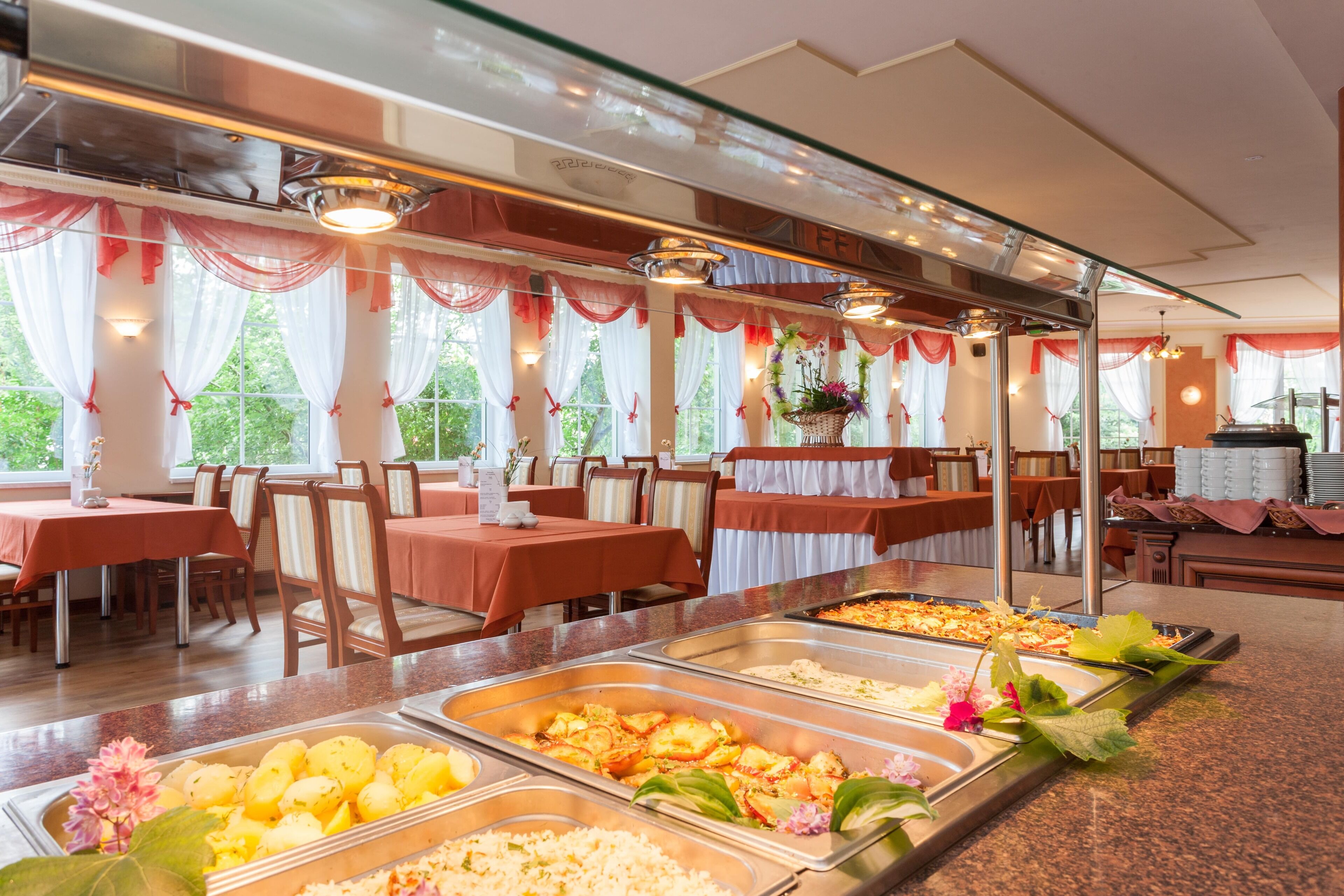 A buffet with various food options, tables with chairs, and large windows with curtains.
