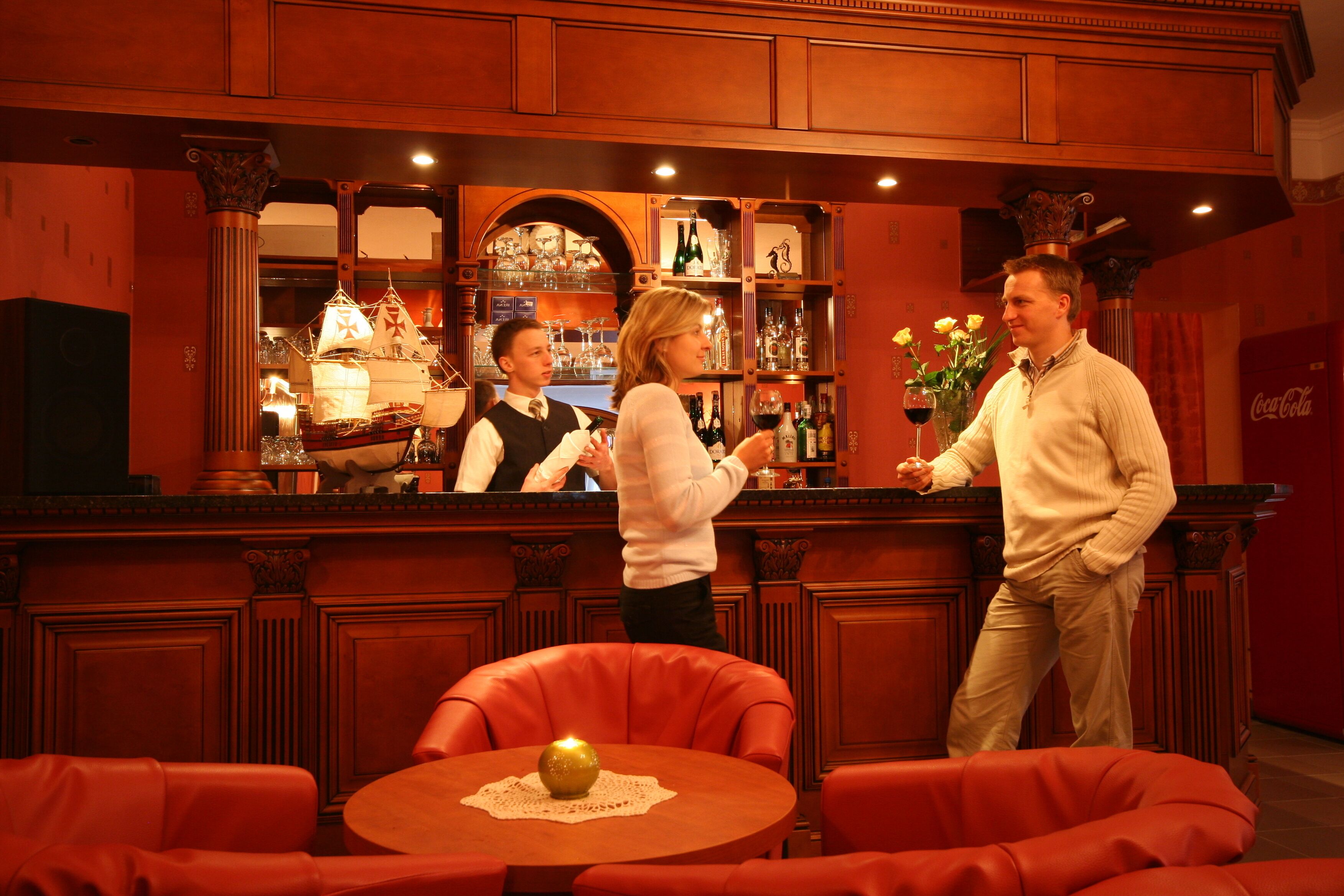 Een bar met houten lambrisering, een toog en een zithoek met rode stoelen.