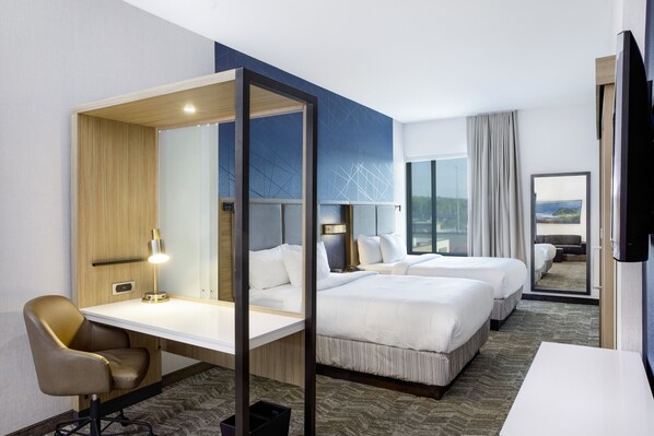 A hotel room with a desk, two beds, a chair, a TV, and a large window with a view.