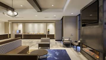 A modern hotel lobby with a seating area, a fireplace, and a kitchen in the background.
