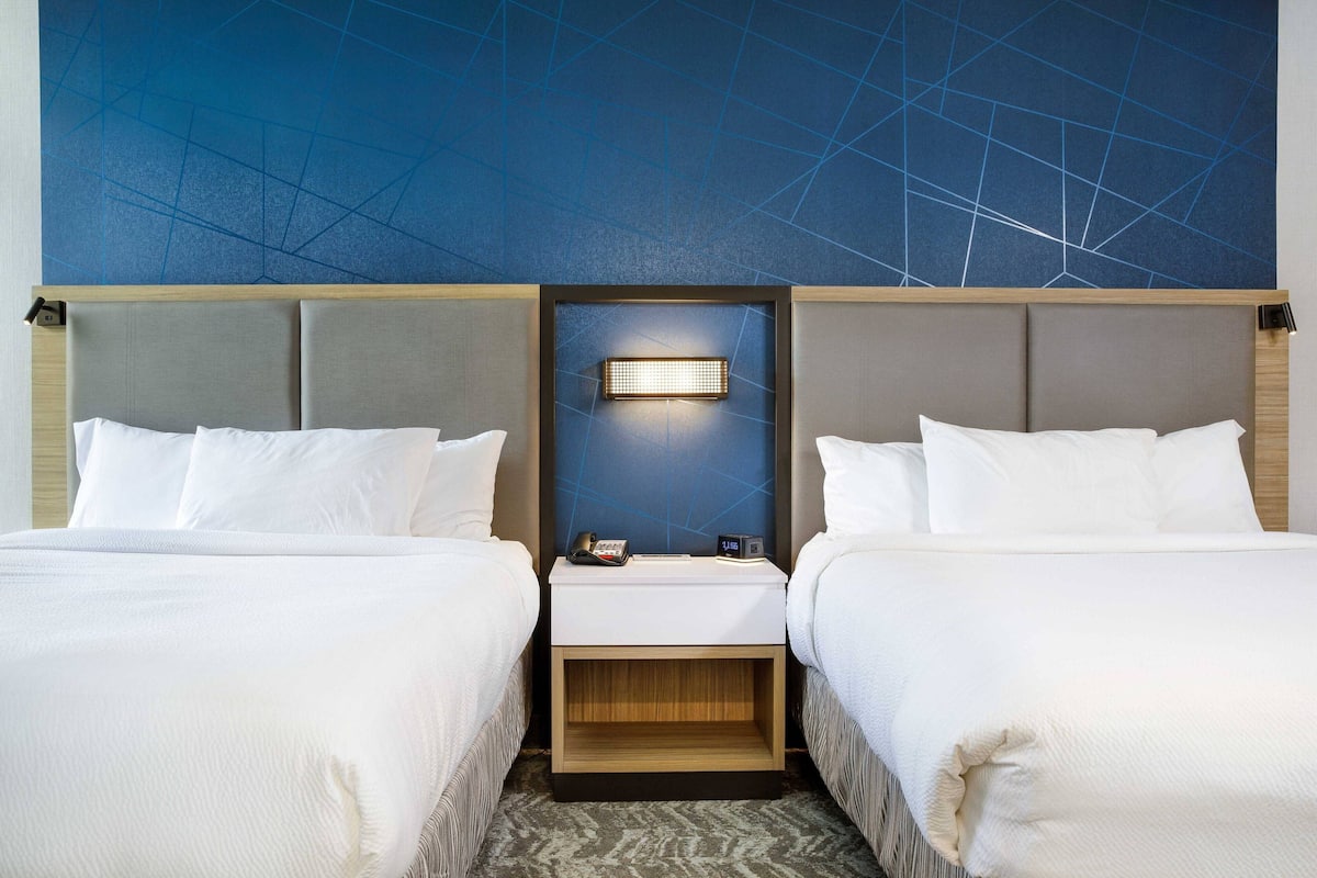 A hotel room with two beds, a nightstand with a lamp, and a geometric wall design.