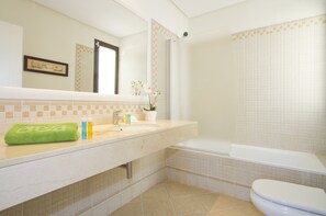 Bathroom features a white bathtub, toilet, and sink with a beige countertop, surrounded by a patterned tile design and a framed picture.