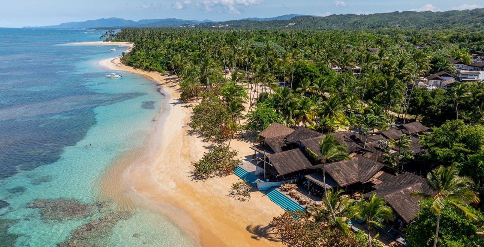 Vue aérienne d’un complexe hôtelier en bord de mer, avec une mer turquoise aux eaux limpides, une plage de sable et plusieurs bungalows aux toits de chaume dotés de piscines.