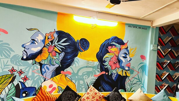 A colorful reception desk with a mural of two women in profile, a yellow wall, and a patterned bookshelf. - Mad Squad Hostel (Varanasi)