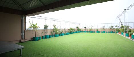 An indoor sports court with artificial turf, surrounded by potted plants and a metal roof structure.