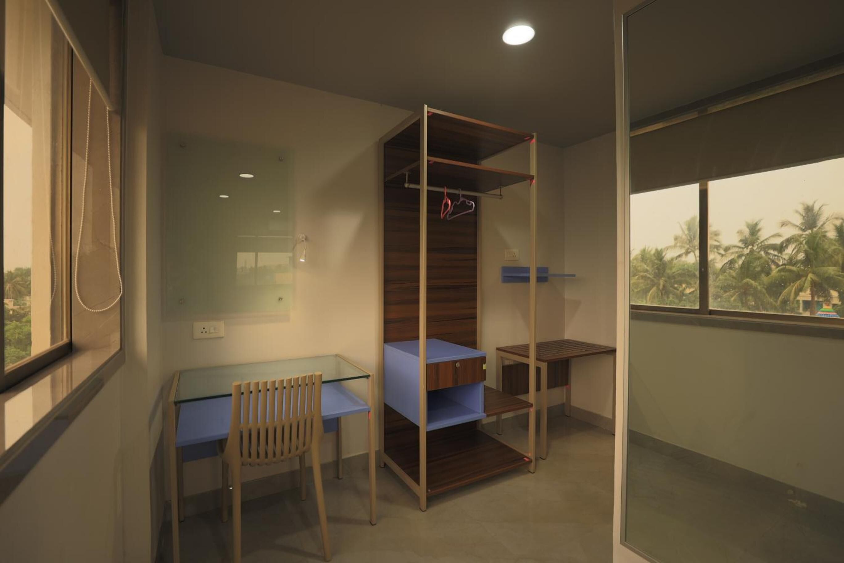 A compact room with a glass desk, a chair, a bed with a wooden frame, and a wardrobe with a mirror.