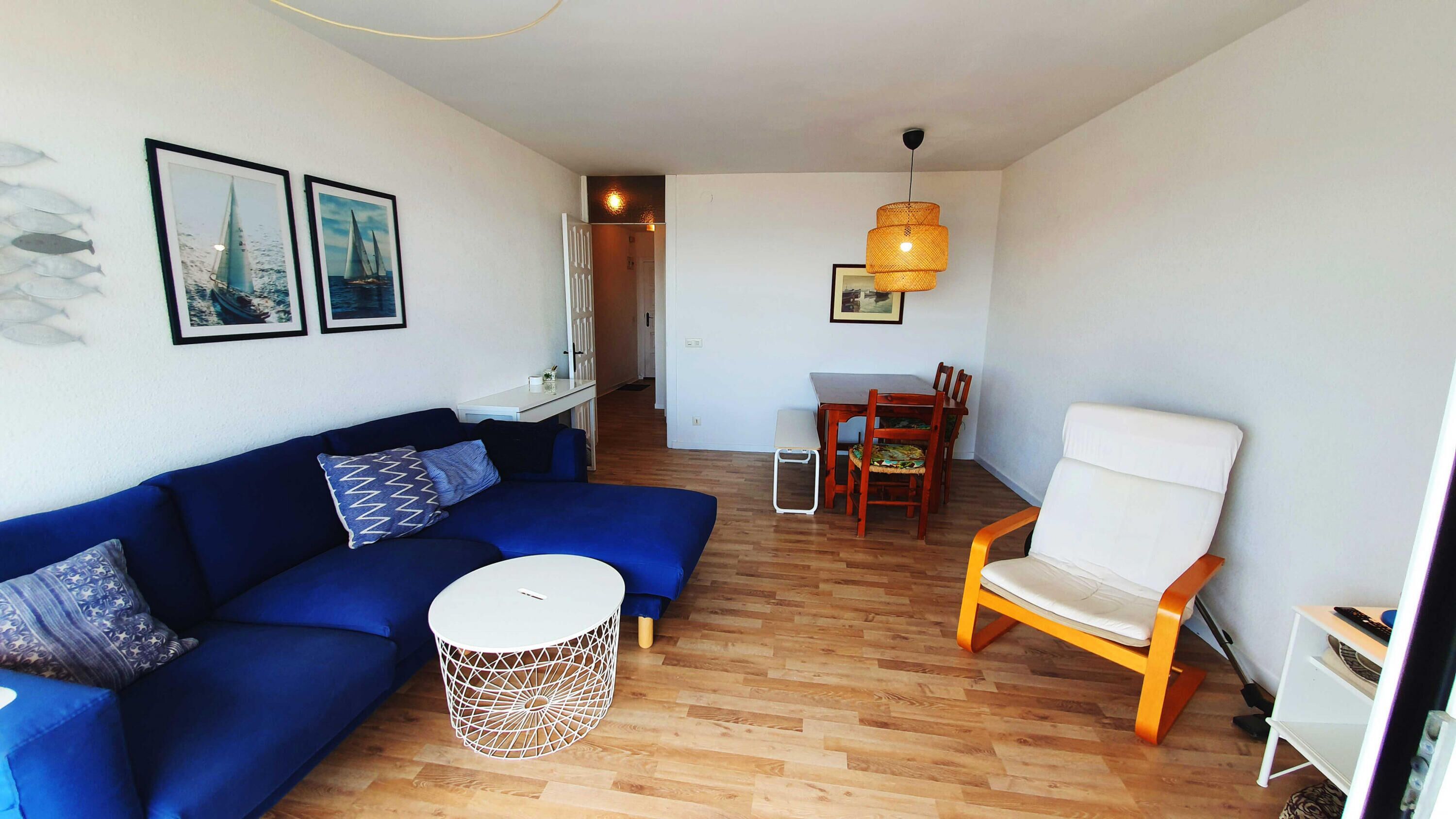 A modern living room with a blue sofa, a white chair, a round coffee table, and a dining area with a wooden table and chairs.