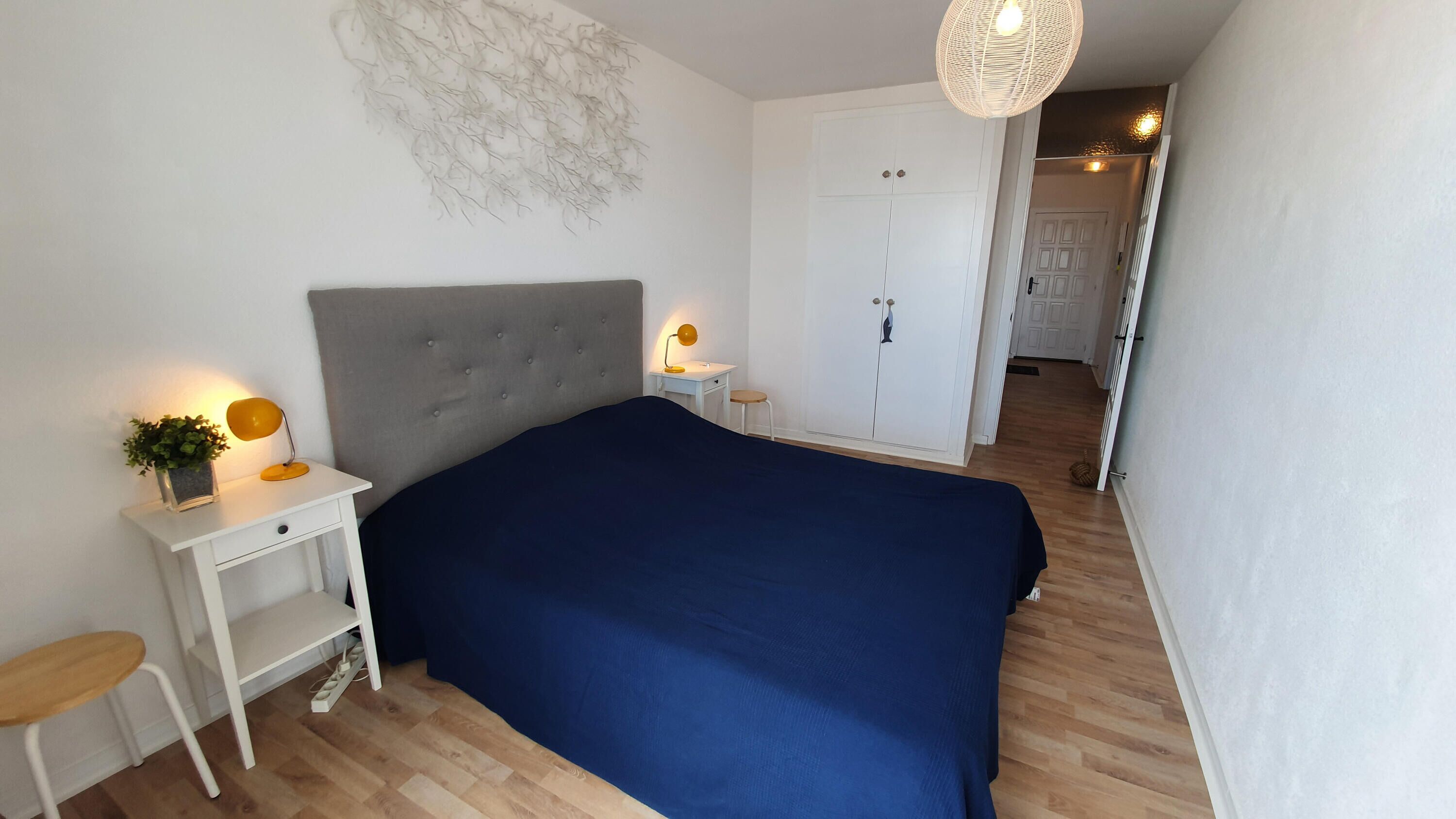 A bedroom with a large bed, a desk with a lamp, a wardrobe, and a hanging light fixture.