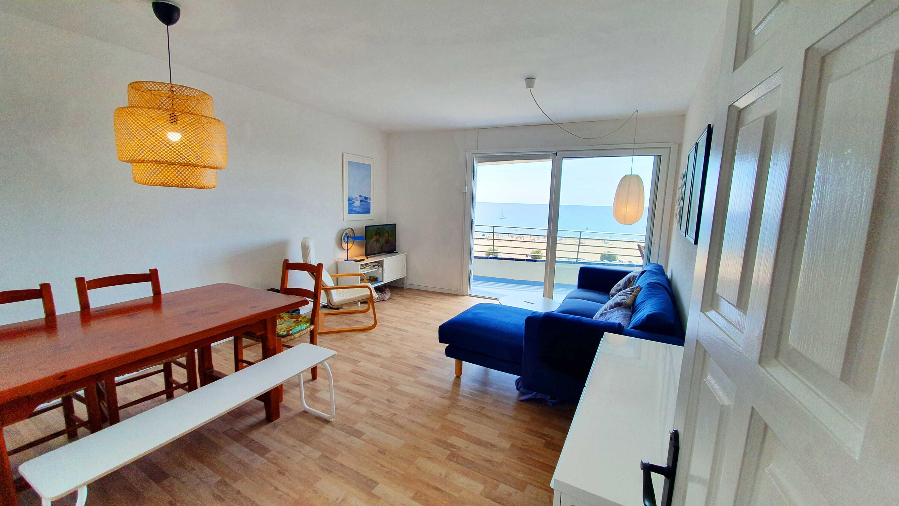 Un salon moderne avec une table à manger en bois, un canapé bleu et un balcon offrant une vue sur la mer.
