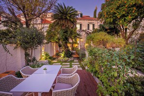 An outdoor seating area with a table and chairs, surrounded by lush greenery and a building with a tiled roof.