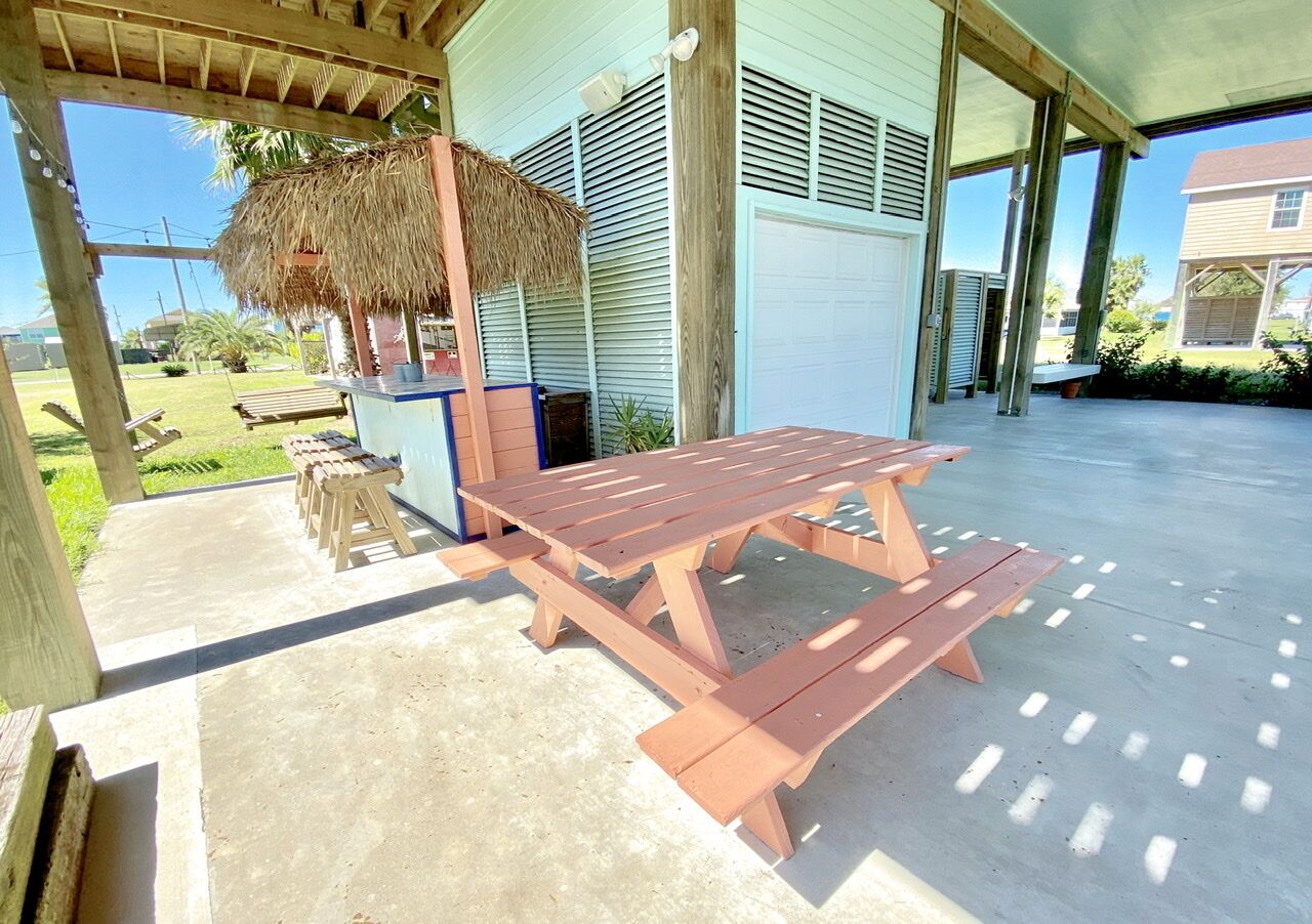A covered outdoor area with a picnic table and benches, a thatched roof structure, and a garage door in the background.