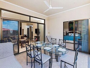 A modern dining area with a glass table and chairs, a ceiling fan, and a view through large glass doors to a living room with a sofa and a TV. - Stay in Tamarindo Condominiums (Tamarindo)