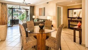 A dining area with a round glass table surrounded by wicker chairs, a wooden side table with a vase, and a view of the outdoors through large windows. - Stay in Tamarindo Condominiums (Tamarindo)