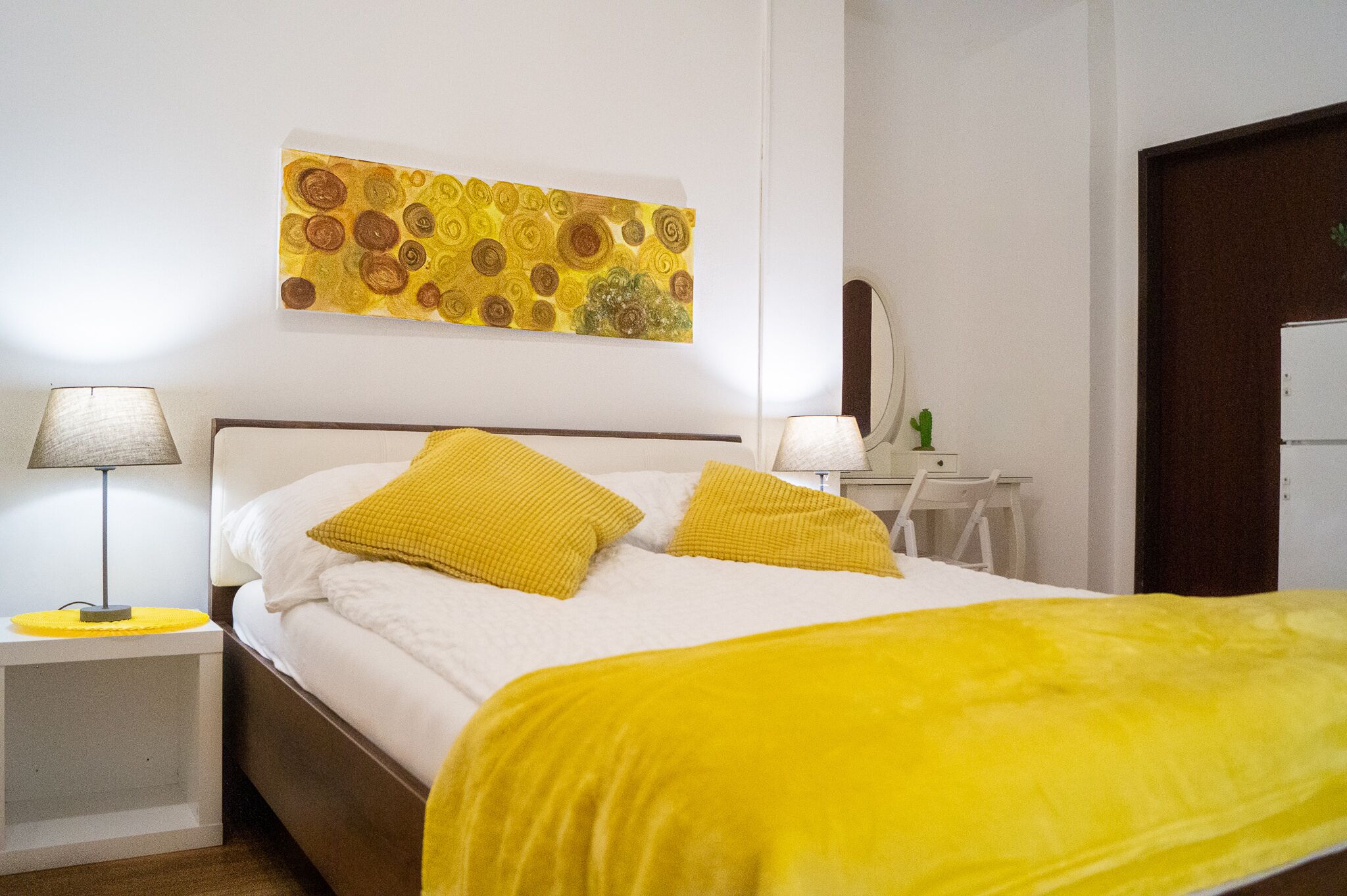 Ein ordentlich gemachtes Bett mit weißer und gelber Bettwäsche, ein Nachttisch mit Lampe und ein Wandbild über dem Bett.