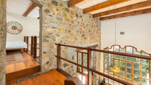 Una habitación rústica con pared de piedra, vigas de madera, una cama, baranda de madera y vistas a través de una ventana.