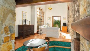 Una habitación con paredes de piedra, una mesa de madera, un banco tapizado de verde, un sofá blanco y un televisor.