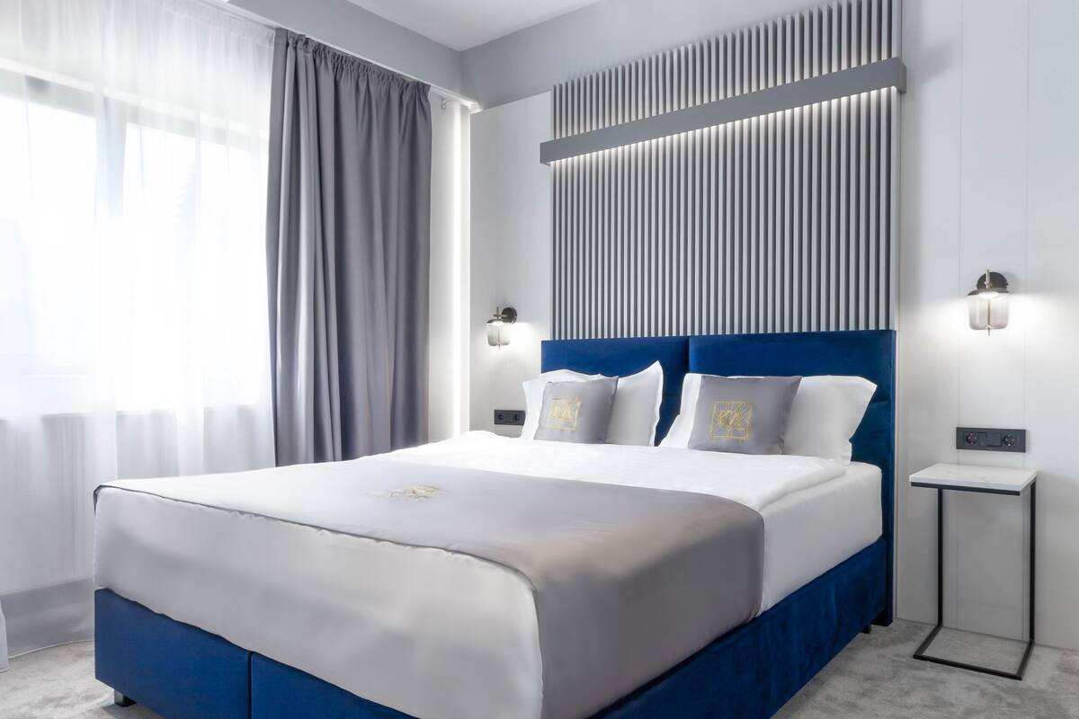 A modern hotel room with a large bed, a headboard with vertical lines, a bedside table with a lamp, and a window with curtains.