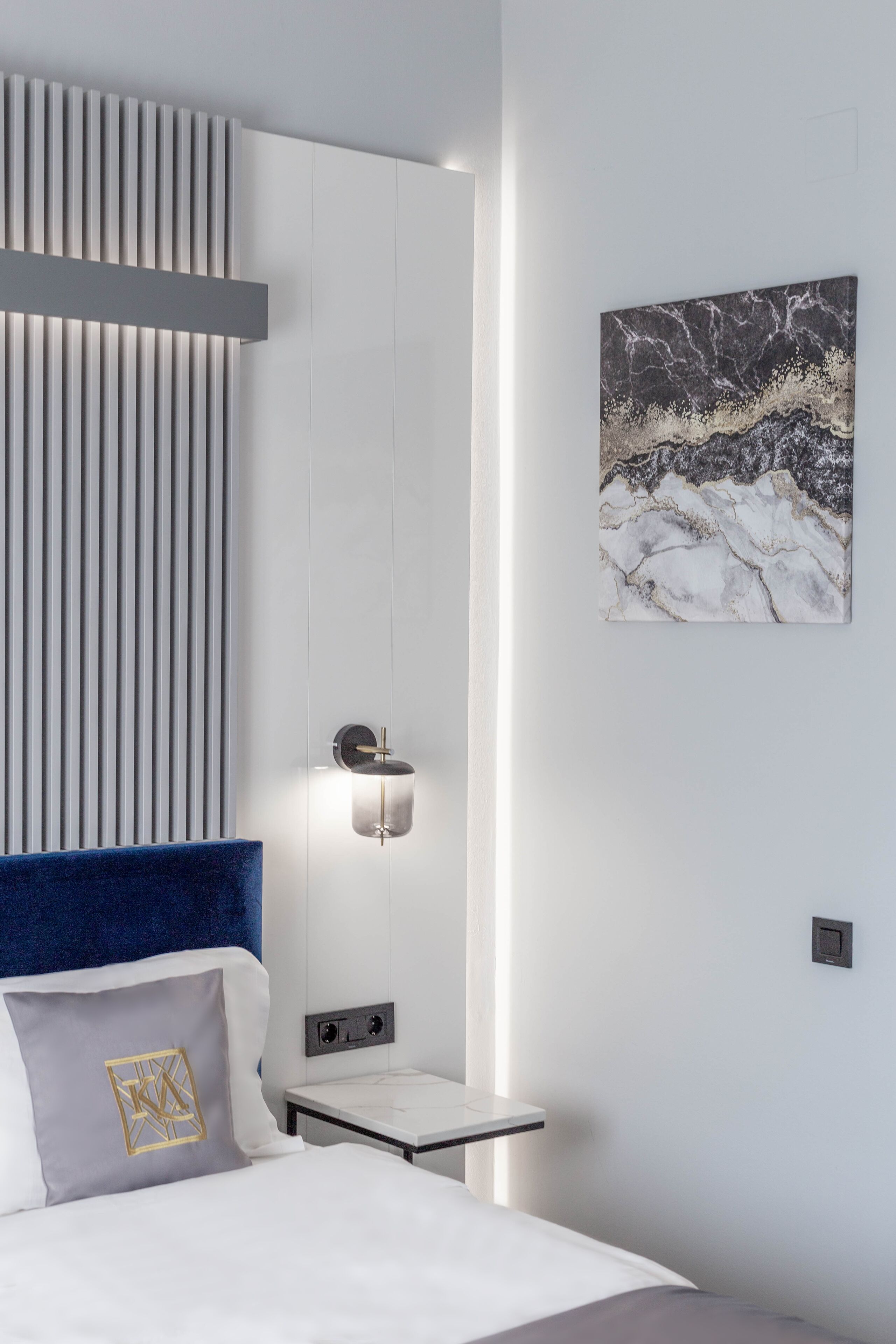 Quarto de hotel moderno com uma cama, uma mesinha de cabeceira, uma luminária fixada na parede e uma obra de arte em mármore.