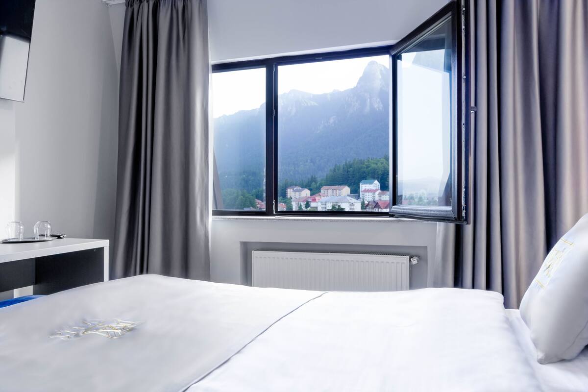 A hotel room with a bed, a desk with a chair, and a window with a view of mountains and buildings.